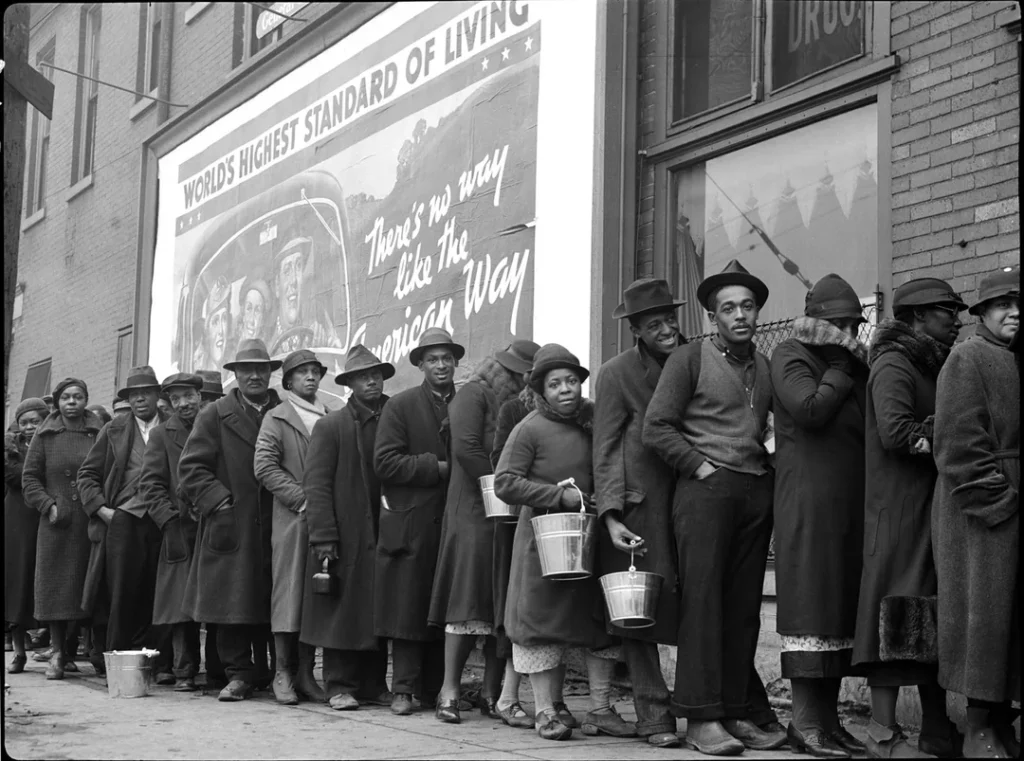 African American flood victims line up to get food and clothing from a Red Cross relief station in front of billboard ironically extolling WORLD'S HIGHEST STANDARD OF LIVING THERE'S NO WAY LIKE THE AMERICAN WAY, taken in 1940