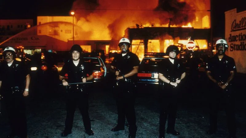 LAPD officers stood guard, as firefighters battled a blaze near 19th Street and Adams Boulevard in Los Angeles during the Rodney King riots. The officers were there to protect the firefighters from being recently shot at by snipers and drive-by-shooters.