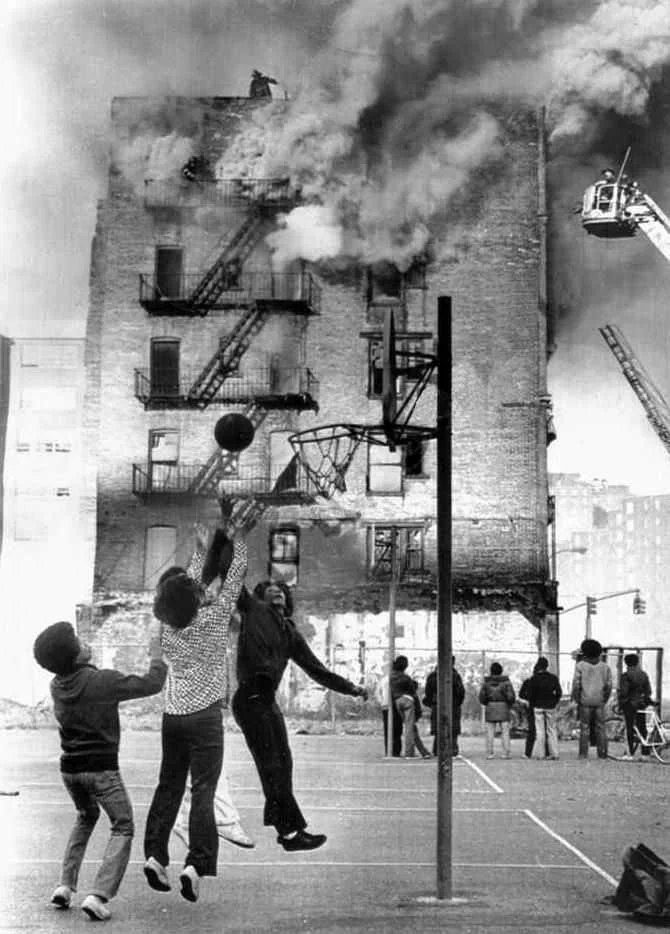 The game goes on while firefighters battle a smoky blaze at a building on 101st Street at First Avenue in East Harlem on Feb. 3, 1975
